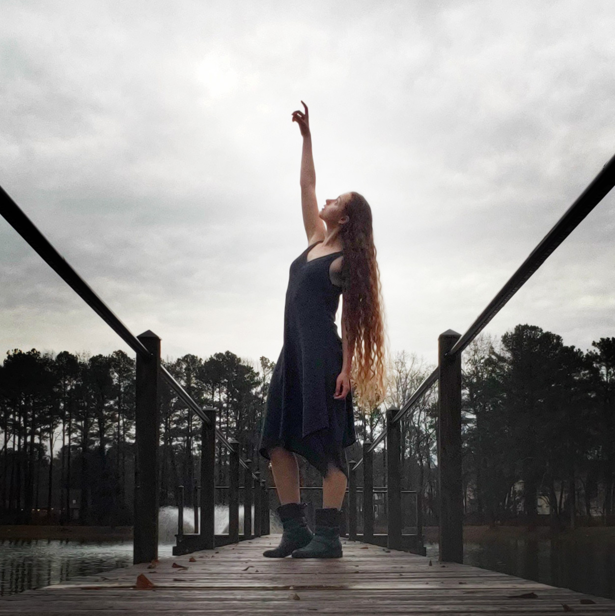 Hananiah points and looks towards upwards dressed in a black dress and black elfish boots on a pier on a lake surrounded by tall dark trees. The ends of her flowing red hair are lit up by the overcast sky behind her